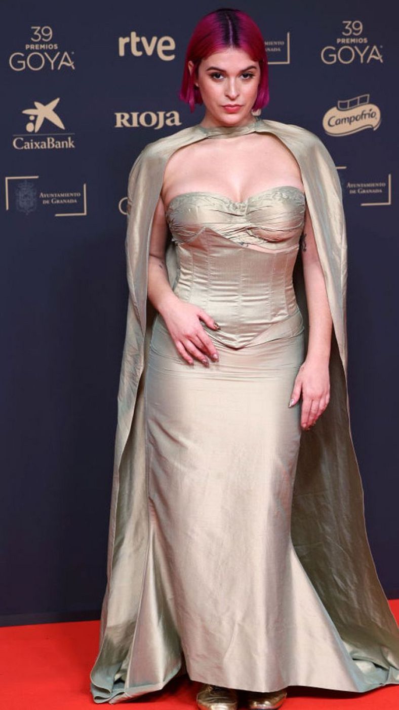 Dora, con vestido beige y capa, posa en la alfombra roja de los Premios Goya.  Pelo corto rosa. Fondo con logotipos de patrocinadores.