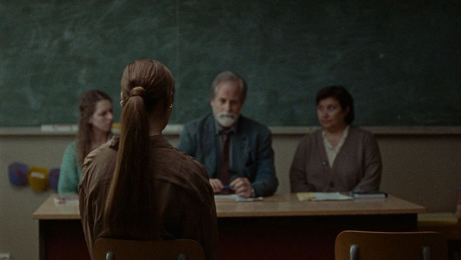 Escena de aula con una estudiante y tres adultos, posiblemente profesores, en una reunión o tutoría.  Pizarra verde al fondo.