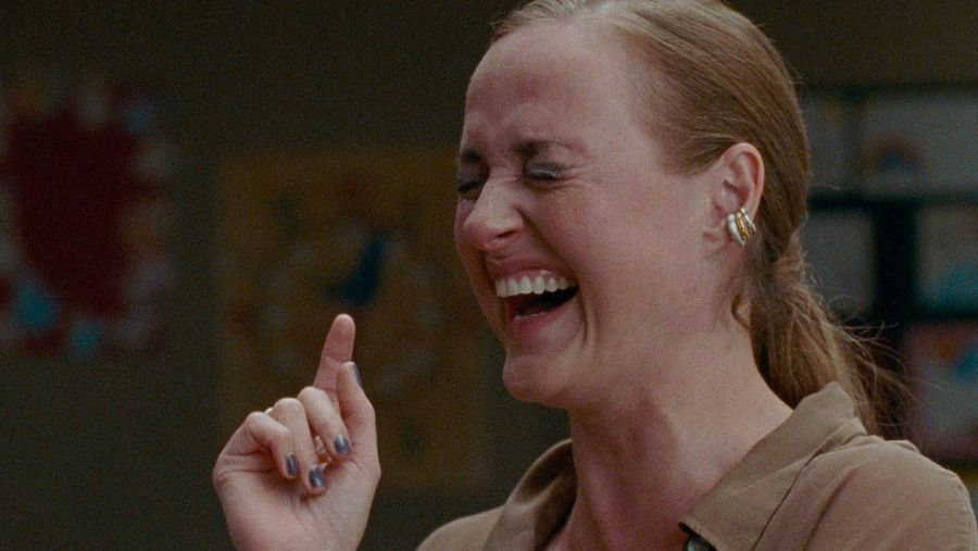 Mujer riendo con intensidad, dedo índice levantado, pelo recogido y camisa marrón claro. Fotograma de interior, posiblemente un aula.