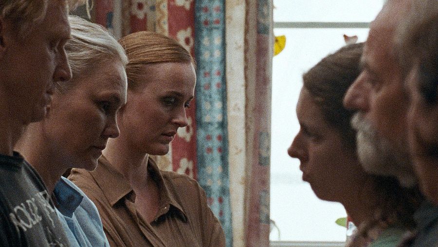 Cuatro personas, dos hombres y dos mujeres, conversan en un interior cerca de una ventana con cortinas rojas y azules. Sus expresiones sugieren tensión.