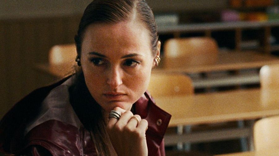 Mujer joven, cabello recogido, chaqueta granate, expresión seria, mano en barbilla con anillos, aula con mesas y sillas de madera.