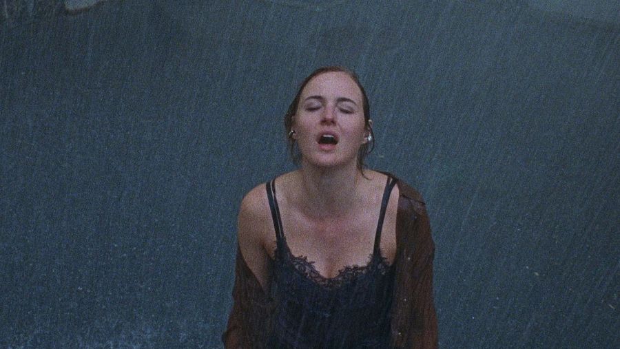 Joven con expresión de angustia bajo lluvia intensa; viste camiseta negra y camisa marrón.  Escena oscura y dramática.