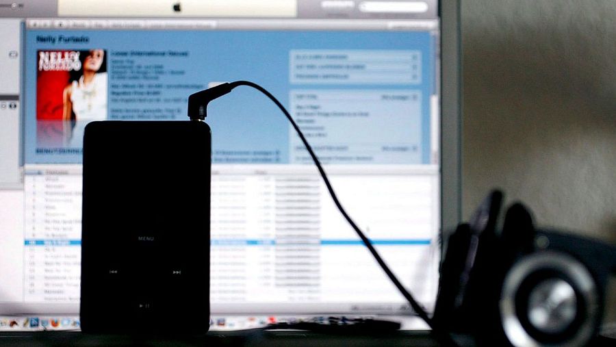 iPod negro conectado a un ordenador mostrando iTunes, con una lista de reproducción y portada de álbum visible. Cámara desenfocada en primer plano.