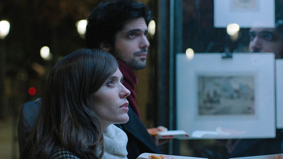 Escena nocturna de dos actores comiendo pizza y mirando hacia arriba a través de un escaparate con cuadros.  Pelo largo y liso en una actriz, pelo rizado y barba en el actor.