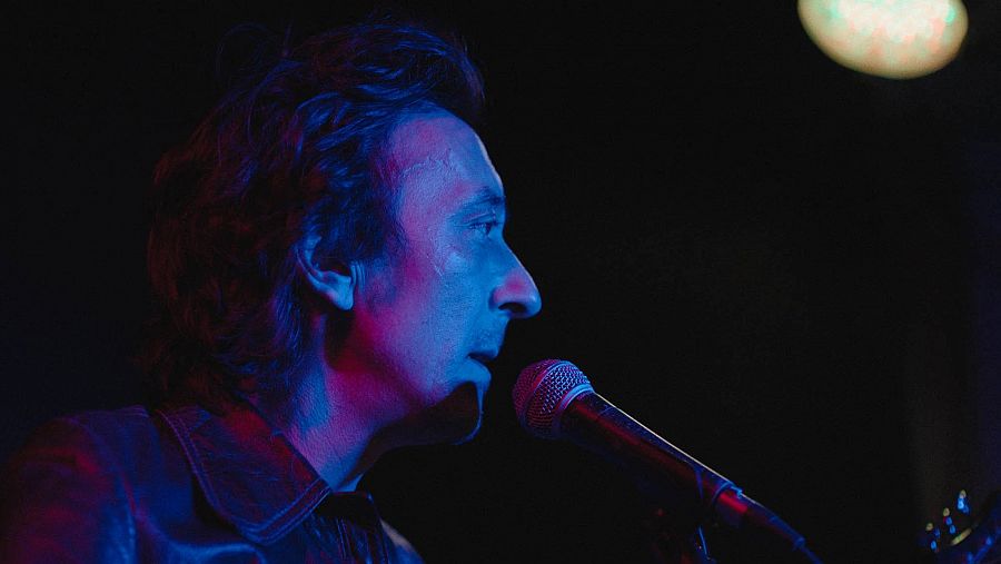 Cantante de perfil sujetando un micrófono durante una actuación con iluminación tenue en tonos azules y rosas.