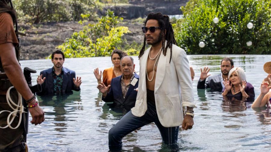 Lenny Kravitz, con rastas y gafas, entra a una piscina en una escena tropical de 'Una boda explosiva'.  Otros presentes muestran sorpresa.