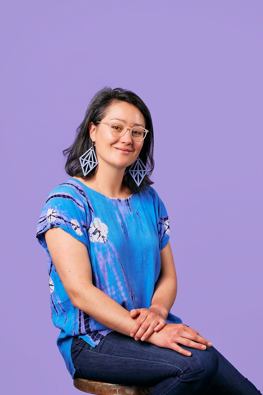 Mujer con gafas y pendientes azules claros, blusa azul tie-dye y vaqueros, sentada sobre un taburete con fondo morado. Mira a cámara con expresión serena.