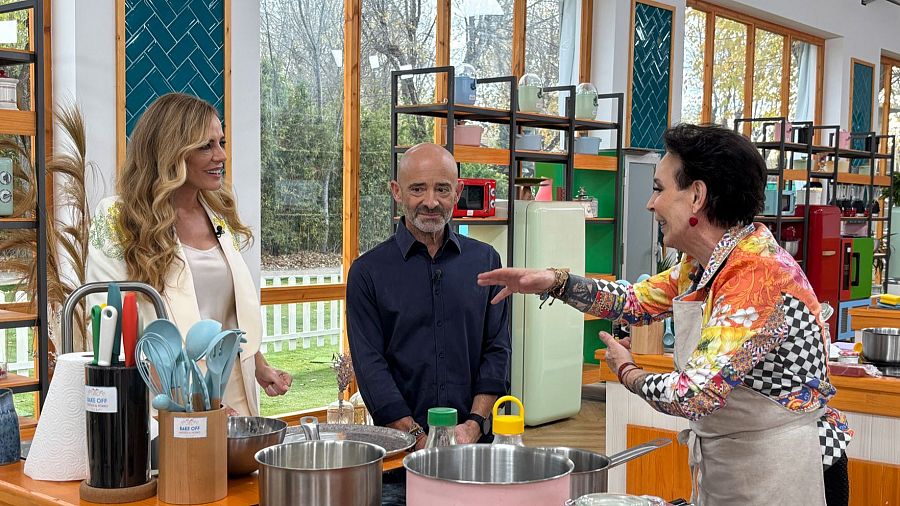 Tres personas en un plató de cocina, con utensilios y recipientes etiquetados 'Bake Off'. Una persona extiende la mano hacia otra, en un ambiente luminoso y colorido.