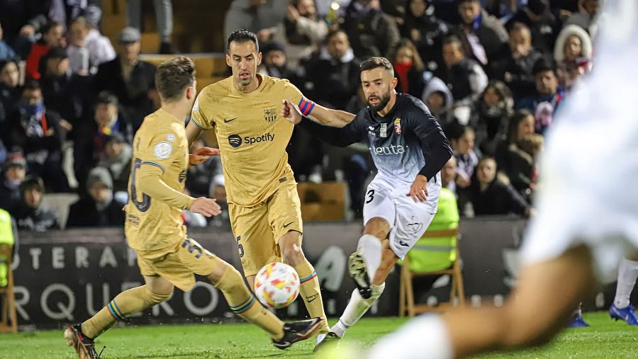 Jugada de fútbol con un jugador del FC Barcelona (camiseta dorada) y otro del AD Ceuta FC (camiseta negra y blanca, dorsal 3) disputando el balón.