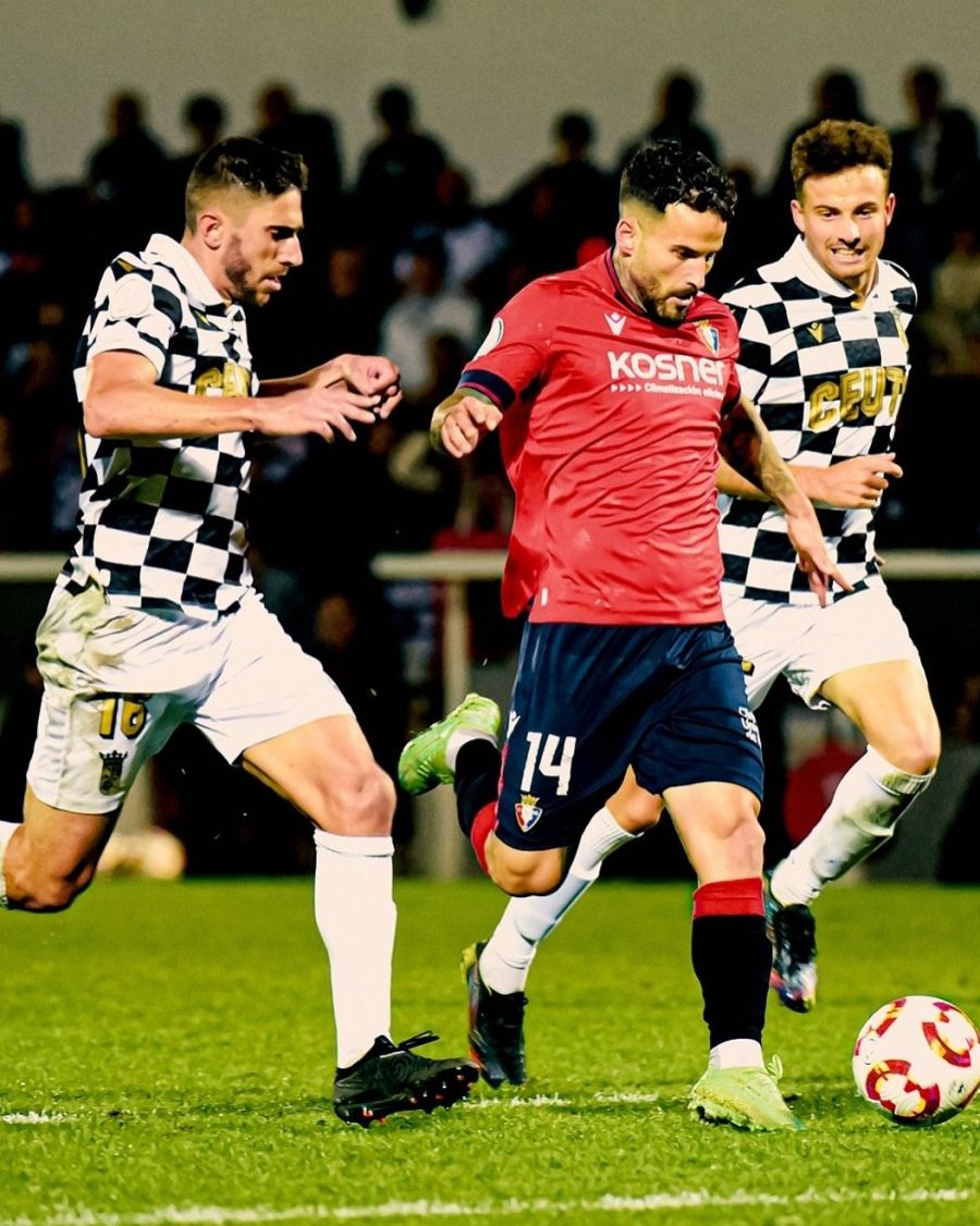 Jugada de fútbol: jugador con camiseta roja y pantalón azul (nº 14) dribla a dos rivales con camisetas blancas y negras (uno con el nº 16). Partido disputado de noche.