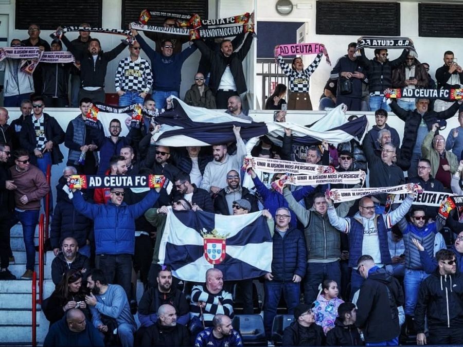Aficionados del AD Ceuta FC en el estadio Alfonso Murube, mostrando su apoyo con bufandas, banderas y pancartas con los colores del equipo y el lema 