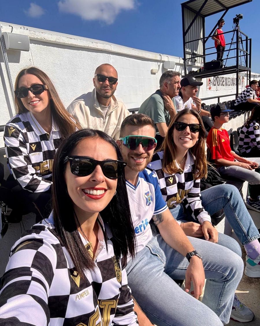 Afición del AD Ceuta FC en el estadio, con camisetas a cuadros blanco y negro y detalles dorados. Dos mujeres con gafas de sol sonríen.