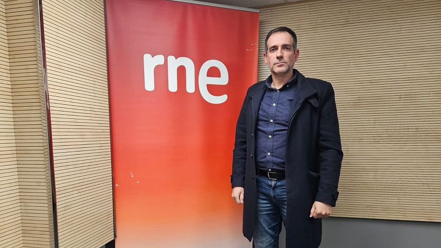 Juan José Liarte, en estudios RNE, posa ante un panel rojo anaranjado con el logo 'rne' en blanco. Viste camisa azul oscura, vaqueros y abrigo.