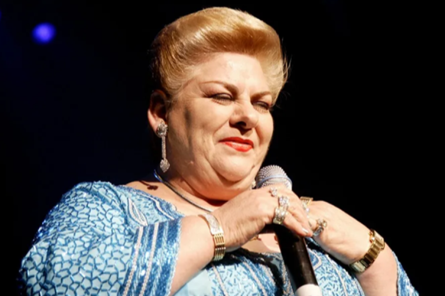 Paquita la del Barrio, cantante mexicana, con vestido azul brillante y micrófono, cabello rubio recogido y expresión serena.