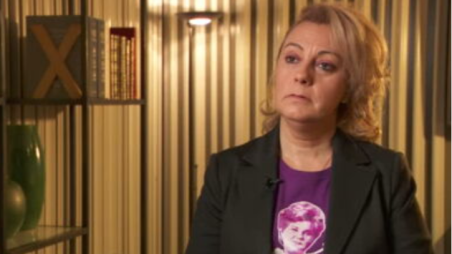 Primer plano de Raquel Orantes en una entrevista; expresión seria, chaqueta negra sobre camiseta morada con diseño. Fondo con estantería.