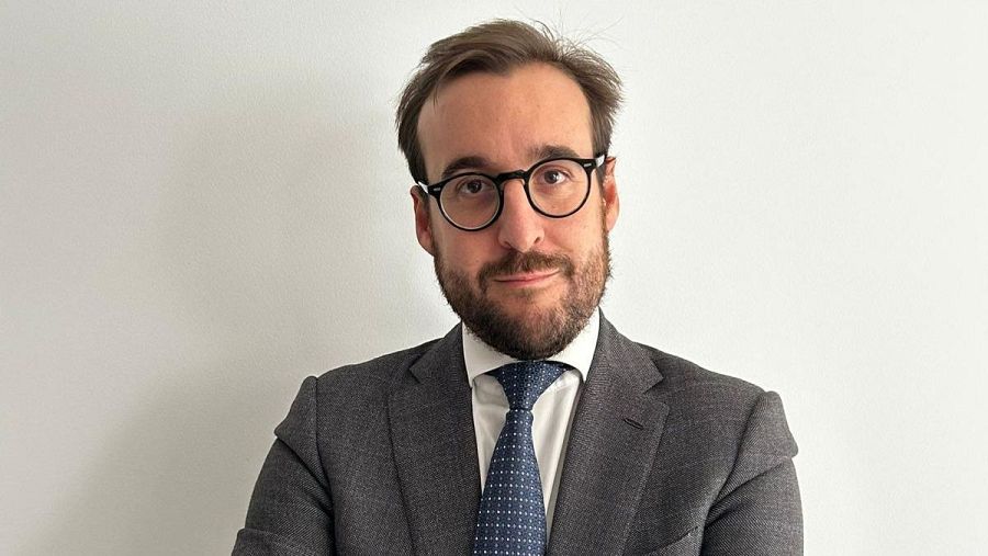 Retrato de un abogado con traje gris, corbata azul marino y gafas, expresión seria, fondo blanco. Imagen profesional.