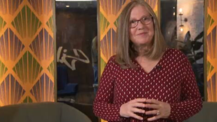 Mujer con blusa granate y gafas, manos juntas, fondo con diseño geométrico naranja y verde.  Contexto: Objetivo Igualdad.