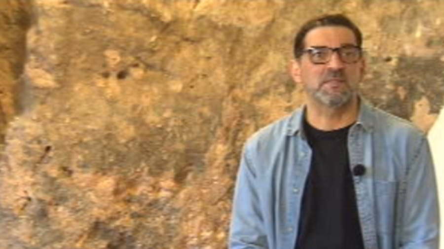 Presentador con camisa vaquera y gafas frente a pared rocosa. Pelo castaño recogido y barba.