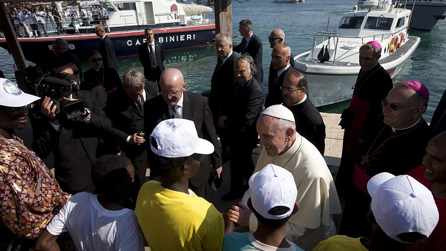 El Papa Francisco conversa con migrantes en Lampedusa, mostrando cercanía.  Se aprecia un joven con camiseta amarilla y otros con gorras blancas.  Al fondo, barcos en el puerto.