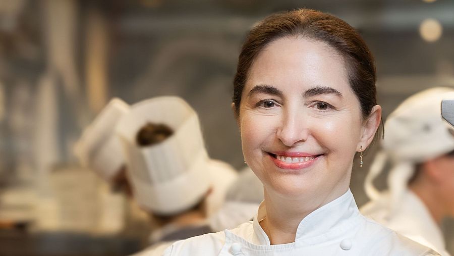 Retrato de Elena Arzak, chef con uniforme blanco, sonriendo. Fondo borroso con ambiente de cocina.