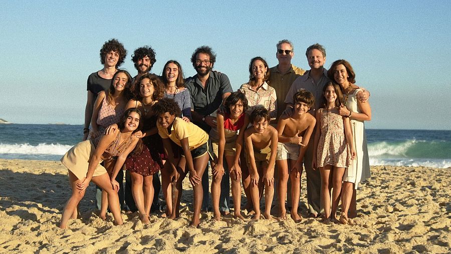 Familia o grupo numeroso en playa, posando para foto.  Ropa estilo años 70.  Ambiente alegre y unido.