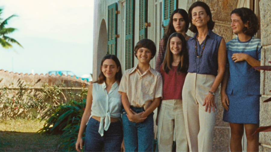 Familia numerosa (7 personas) posando ante casa colonial con persianas verdes. Ropa informal años 70. Madre con blusa azul marino y pantalón beige.
