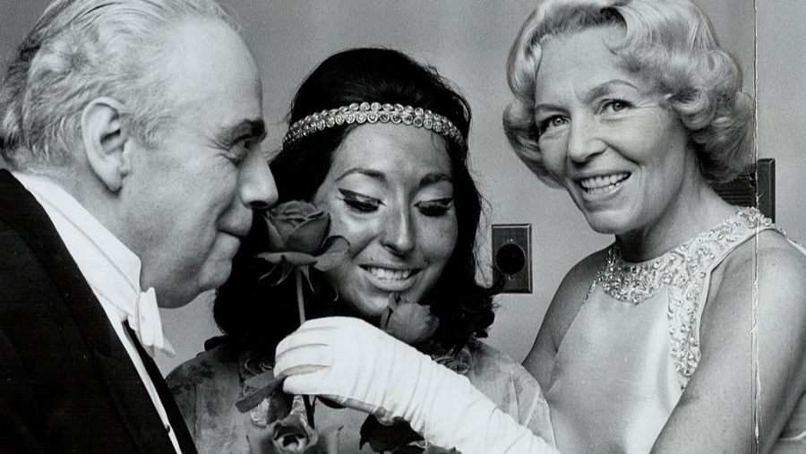 Tres personas en una escena formal: un hombre mayor con traje y pajarita, una mujer mayor y una joven con tocado, recibiendo un ramo de rosas.