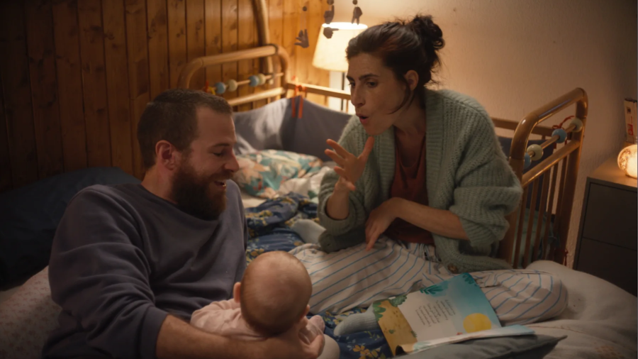 Escena íntima de Álvaro Cervantes y Miriam Galo con un bebé en un dormitorio.  Ella viste un cárdigan verde y pantalones a rayas; él, un jersey azul oscuro.  Ambiente familiar y cálido.