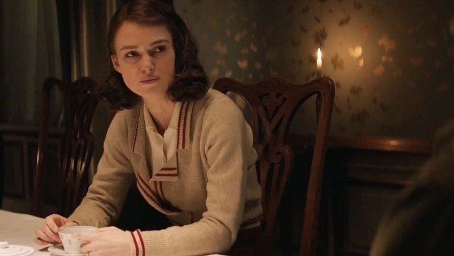 Keira Knightley, con cárdigan beige y expresión pensativa, sostiene una taza de té en una mesa de madera oscura, en una escena íntima iluminada por una vela.