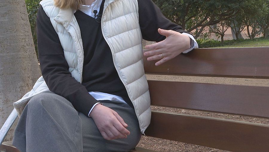 Reportera en entrevista, sentada en un banco de parque. Viste chaleco blanco, jersey oscuro y pantalones grises; usa micrófono de solapa y gesticula con naturalidad.