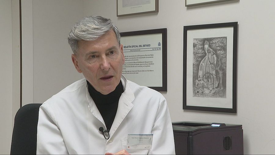 Psiquiatra en su consulta, con bata blanca, frente a un cuadro y un documento enmarcado en la pared.  40 años de experiencia en tratamiento de adicciones.