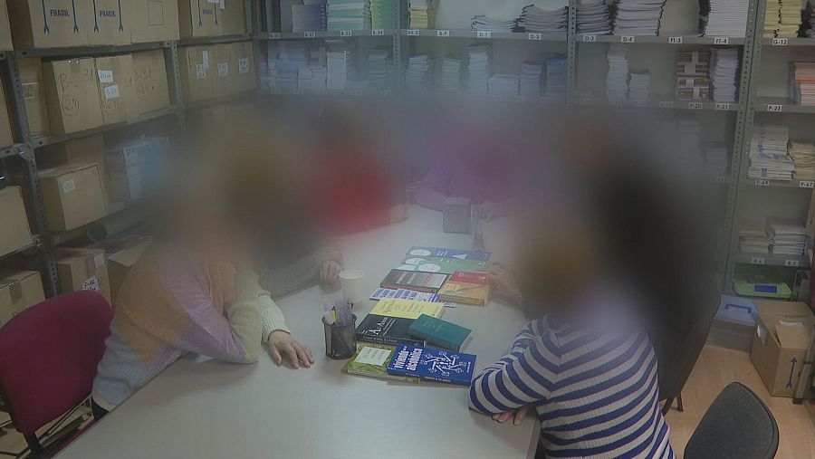 Reunión de grupo con participantes anónimos alrededor de una mesa con libros y folletos.  Se observa material adicional en estanterías.  Ambiente de apoyo y confidencialidad.
