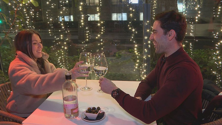Dos personas brindan en un restaurante.  Ella lleva chaqueta beige, él camisa granate.  Sobre la mesa hay copas, agua y aceitunas.