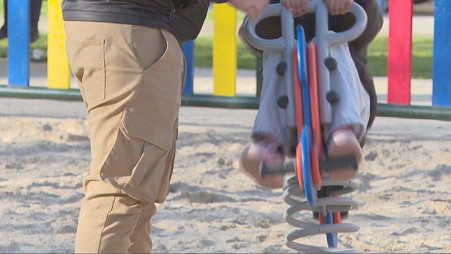 Adulto supervisando a un niño en un balancín de muelle de colores en un parque infantil con suelo de arena.
