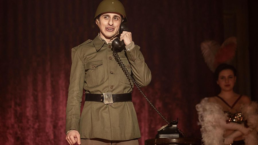 Hombre en uniforme militar hablando por teléfono antiguo en un escenario teatral con fondo rojo.  Mujer con boa de plumas al fondo.