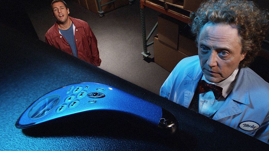 Adam Sandler, sorprendido, observa un mando a distancia azul. Al fondo, un hombre con bata y pajarita mira con seriedad. Escena de la película *Click*.