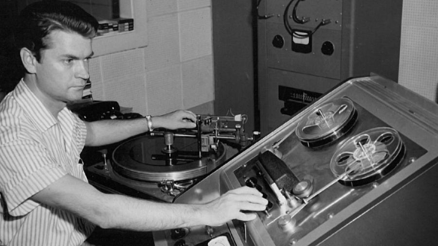 Sam Phillips, en blanco y negro, ajusta un tocadiscos y una grabadora de cinta, posiblemente durante una sesión de grabación musical en un estudio.