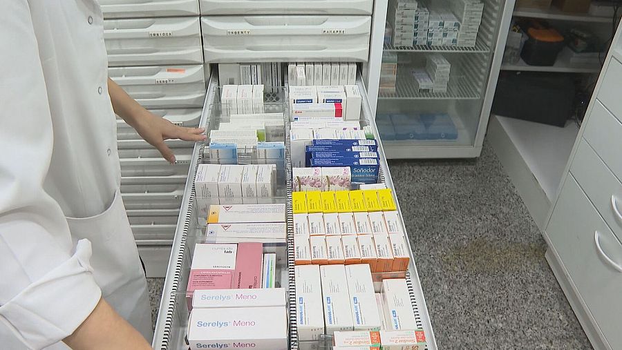 Cajón de farmacia con medicamentos (Serelys Meno, Solfeidar visibles) abierto por un farmacéutico con bata blanca.  Se ve un refrigerador cercano.