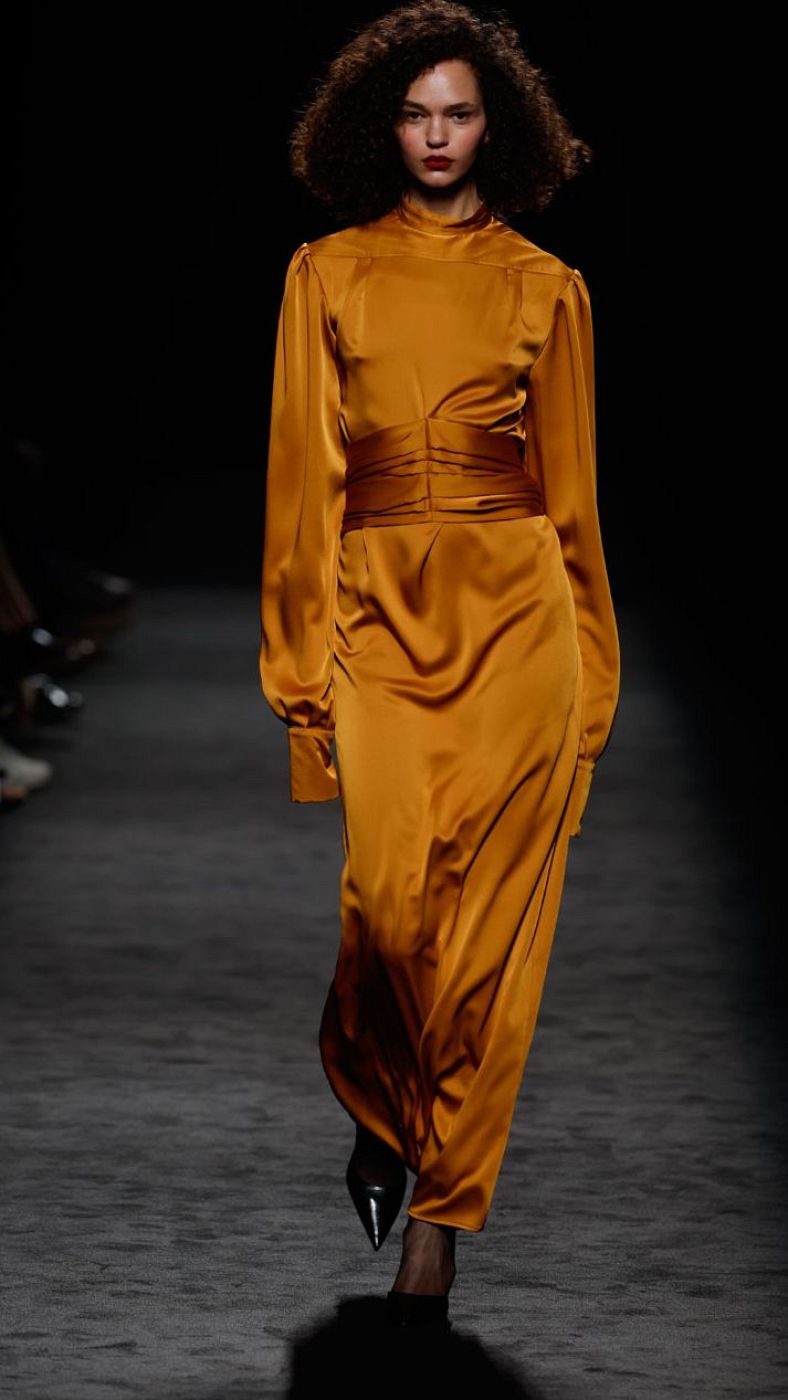 Modelo en pasarela oscura con vestido largo dorado, mangas abullonadas y cuello alto, cinturón fruncido. Zapatos negros de tacón. Público en penumbra.