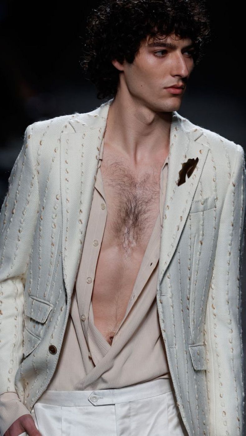Modelo masculino luce americana blanca con detalles cosidos, camisa beige y pantalón blanco en un desfile de moda. Broche oscuro en la solapa.