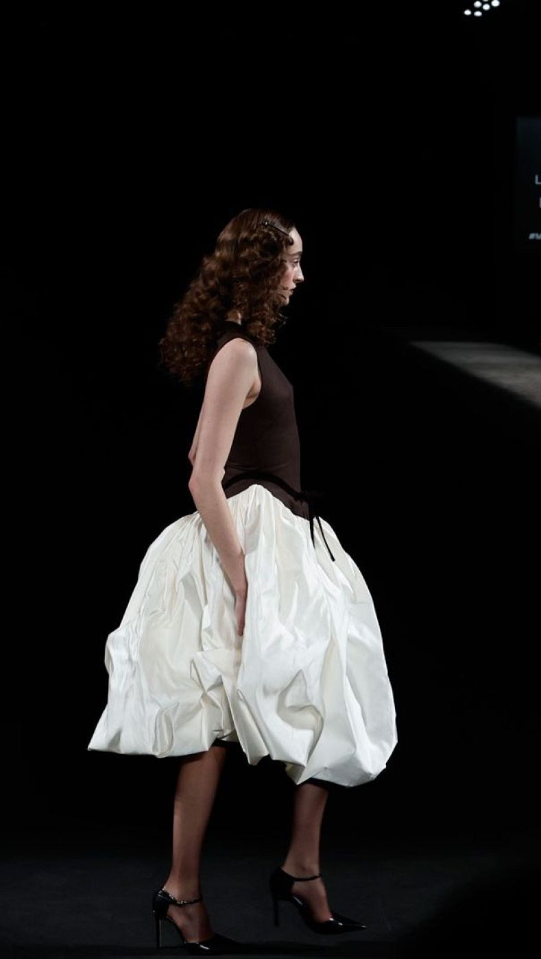 Modelo con vestido marrón oscuro y falda blanca amplia desfila en pasarela oscura, con medias oscuras y zapatos negros de tacón.