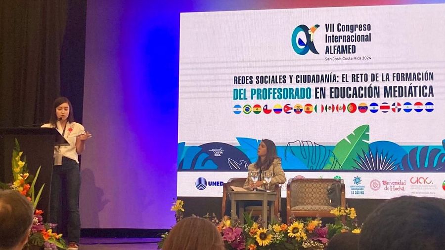 Periodista presentando investigación en congreso internacional.  Se observa un atril, micrófono, pantalla con logotipos y público.