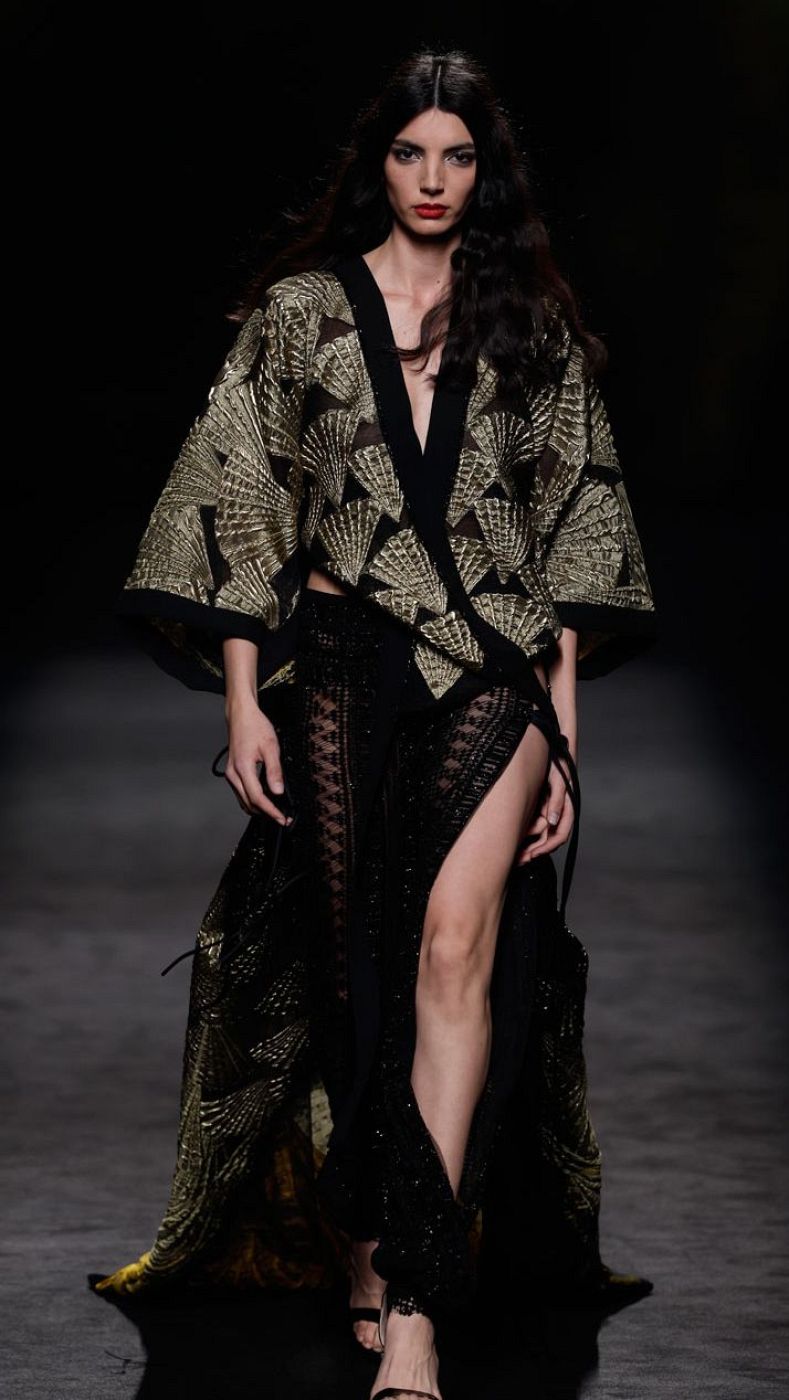 Modelo en pasarela con kimono estampado dorado y falda negra con aberturas, sandalias altas y cabello suelto.