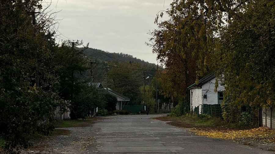 Calle de un pueblo con casas, árboles otoñales y cielo nublado.  El asfalto muestra deterioro.  La imagen no muestra signos de conflicto.
