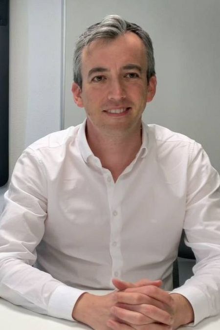 Hombre de mediana edad, cabello canoso corto, camisa blanca formal, sentado con las manos entrelazadas, leve sonrisa.