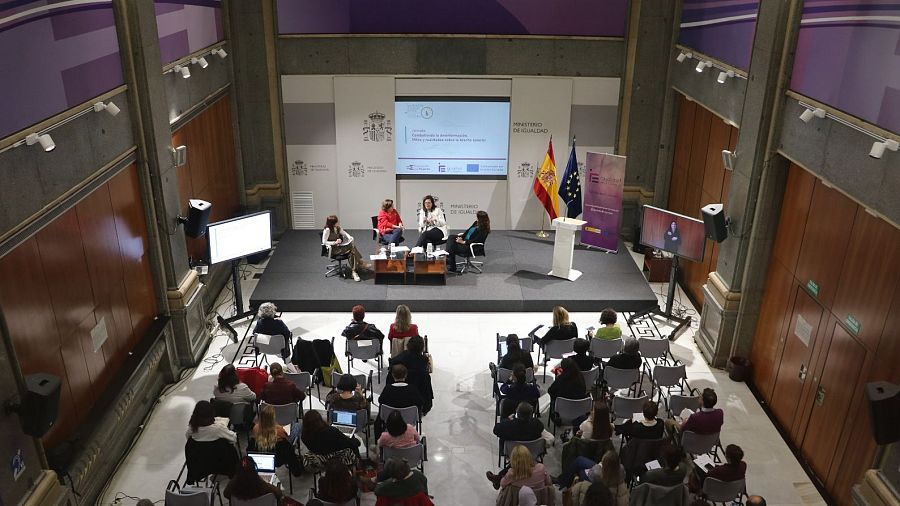 Auditorio con mujeres en una mesa redonda sobre igualdad de género.  Público mayoritariamente femenino.  Pantalla con presentación.