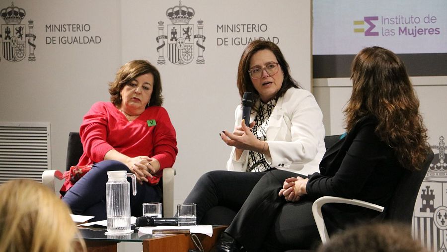 Tres mujeres sentadas en sillas durante una conferencia, una de ellas habla con un micrófono.  Se observan logotipos del Ministerio de Igualdad y el Instituto de la Mujer.