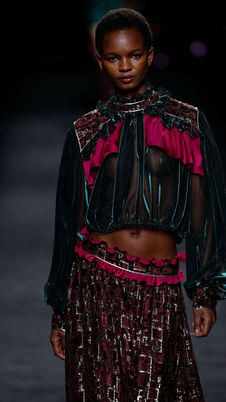 Modelo desfila con top corto semitransparente verde oscuro, detalles fucsia y dorados, y falda larga con estampado geométrico burdeos y dorado.