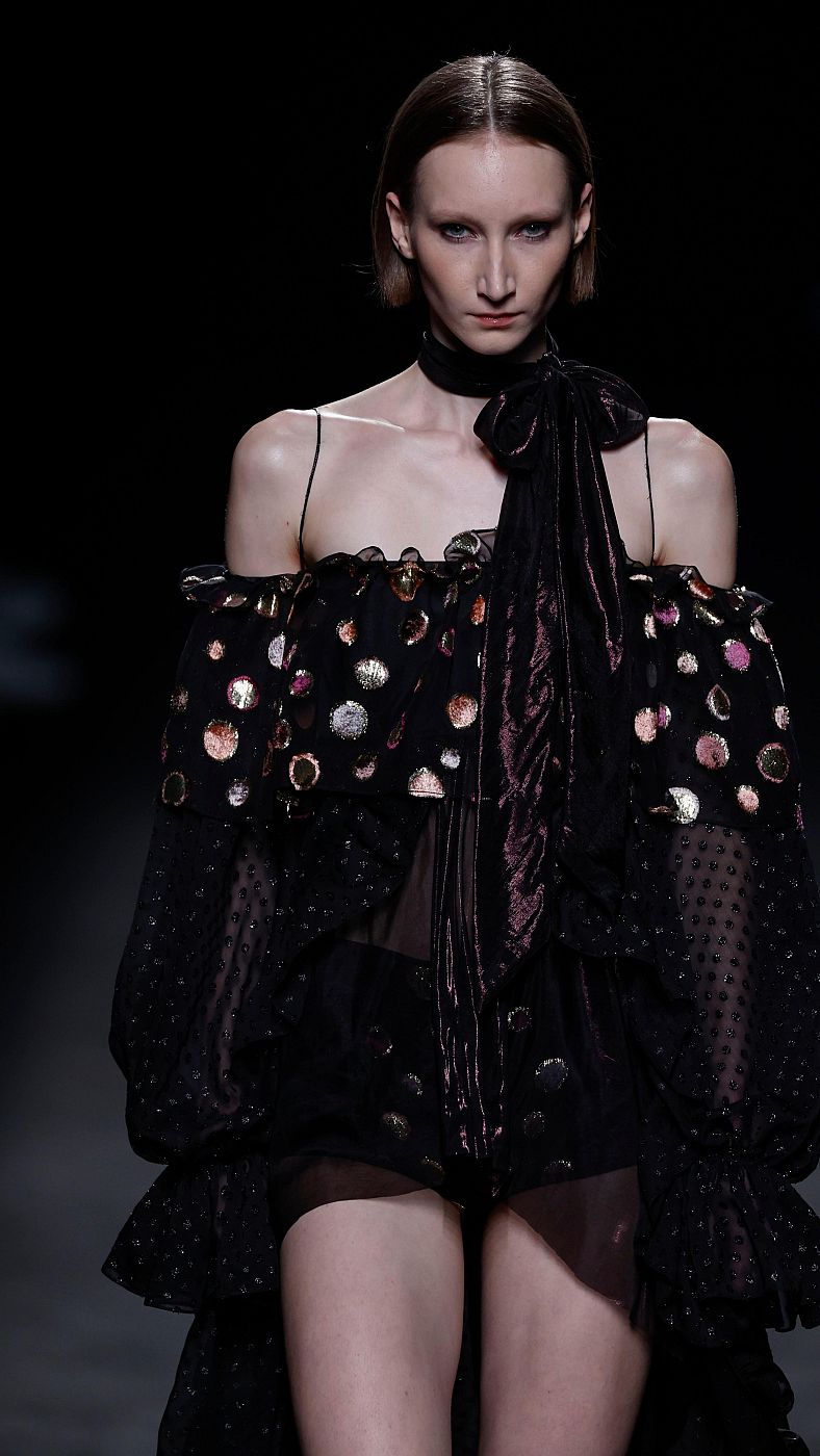 Modelo con vestido negro con volantes, lunares brillantes y lazada granate desfila en pasarela con el pelo recogido.