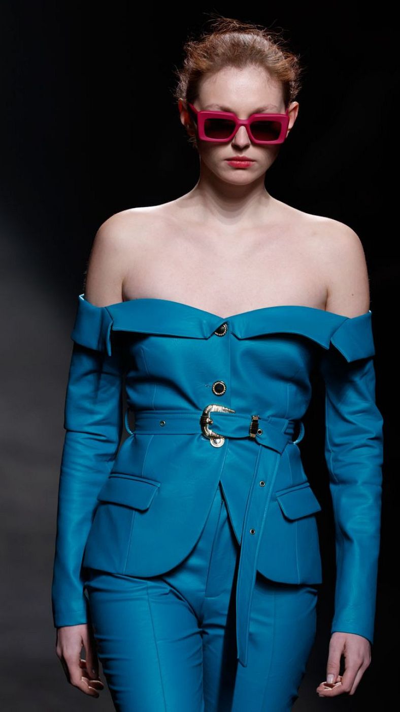Modelo con traje azul turquesa, cinturón dorado y gafas rosas desfilando.  Pelo recogido. Fondo oscuro.